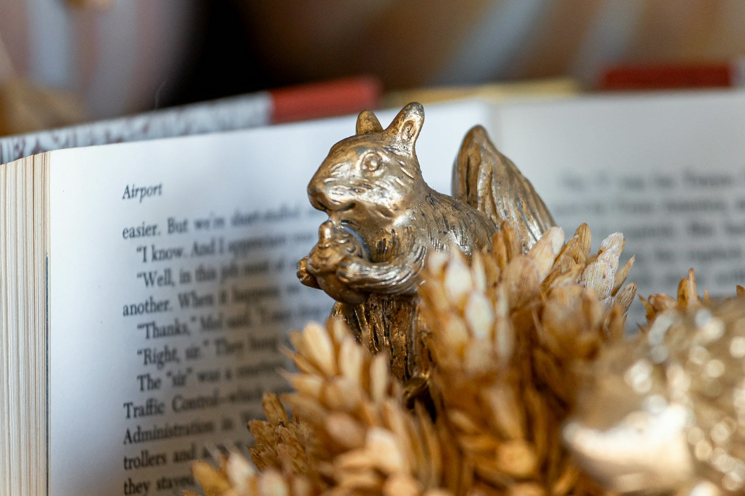 A small metallic squirrel figurine perched on an open book, surrounded by dried pinecones and autumn-themed decorative elements in a warm, shallow depth-of-field setting.